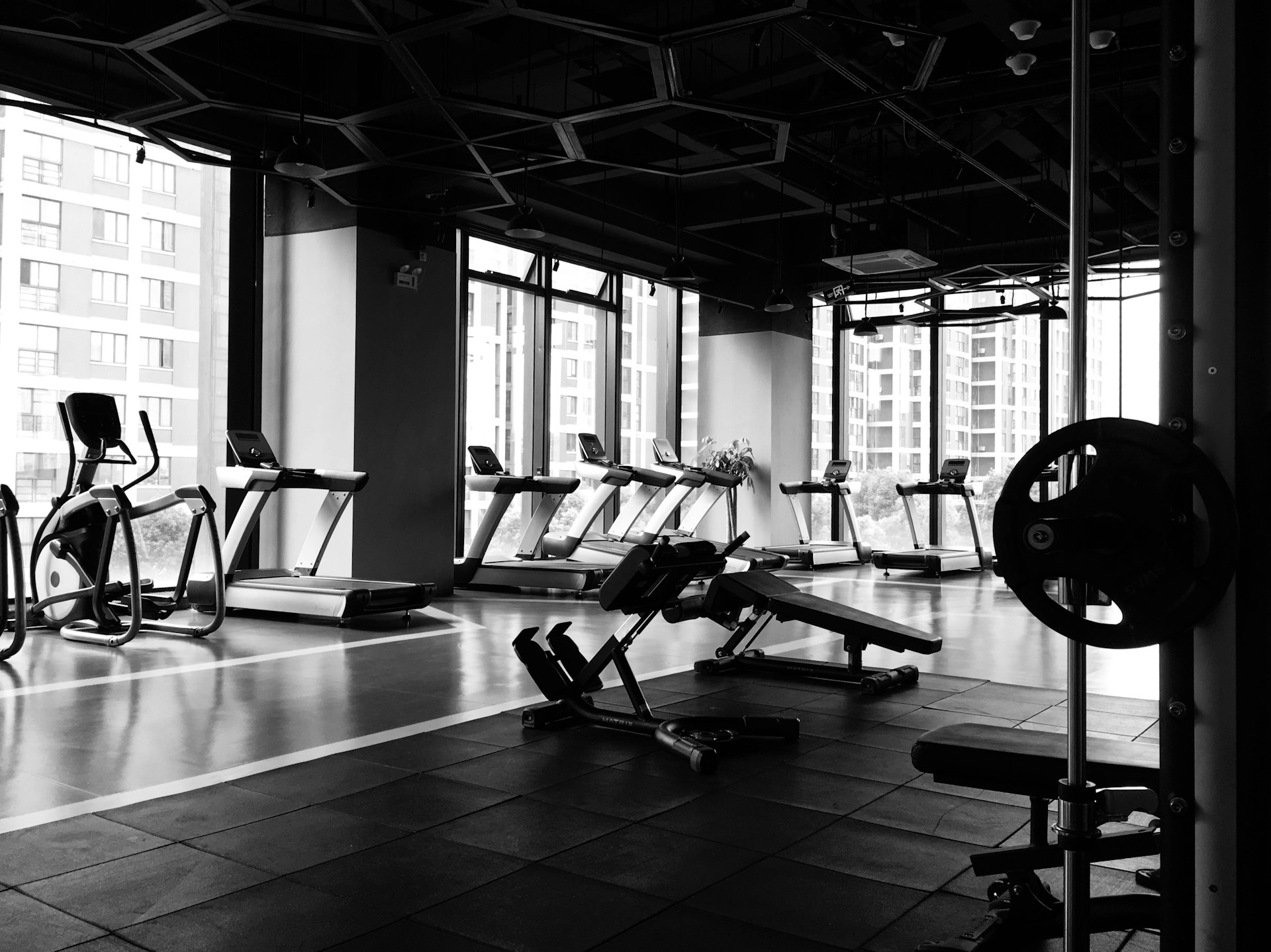 Black and white photo of a gym interior. Large windows, treadmills, and lifting equipment are shown.